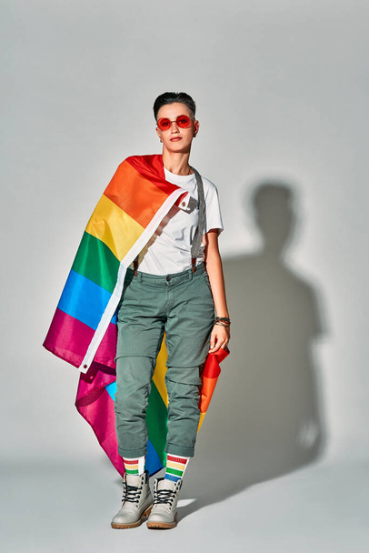 Happy beautiful woman in red eyewear smiling while posing with rainbow flag isolated over white background. Vertical photo. - Photo, Image