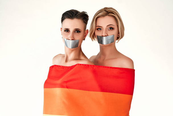 虹の旗に包まれた口テープで検閲された2人の女性。LGBTの権利、検閲概念。隔離された白い背景. - 写真・画像
