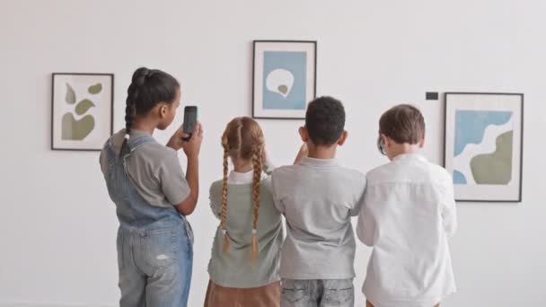 Vista posteriore media lunga di quattro studenti delle scuole medie che scattano foto di dipinti a parete nella galleria d'arte, utilizzando smartphone, poi a piedi - Filmati, video