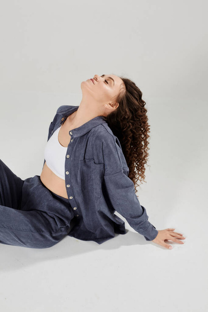 Slim toned model woman in casual clothes is sitting on the floor over white background. Charming young girl with curly hair dressed in a denim costume and white top posing against a white background - Photo, image