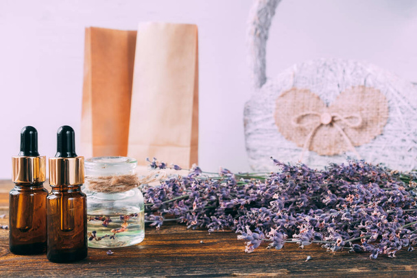 Two bottles of lavender essential oil. On a wooden table against the background of a bouquet of lavender. In the background are small paper bags and a basket. - Foto, Imagen