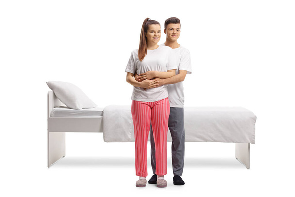 Young couple in pajamas standing in embrace in front of a bed isolated on white background - Fotoğraf, Görsel