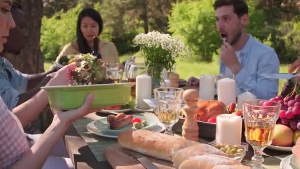 Monietninen ryhmä nuoria mies- ja naispuolisia ystäviä, jotka istuvat yhdessä ruokapöydässä nauttimassa herkullista lounasta kaupungin puistossa lämpimänä kesäpäivänä - Materiaali, video