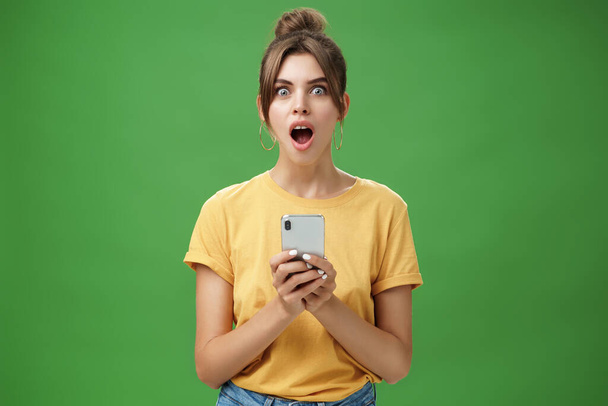 Portret van geschokt sprakeloos en onder de indruk mooi wit meisje met gekamd haar in geel t-shirt met smartphone, dalende kaak van opwinding reageren op koele app over groene achtergrond - Foto, afbeelding