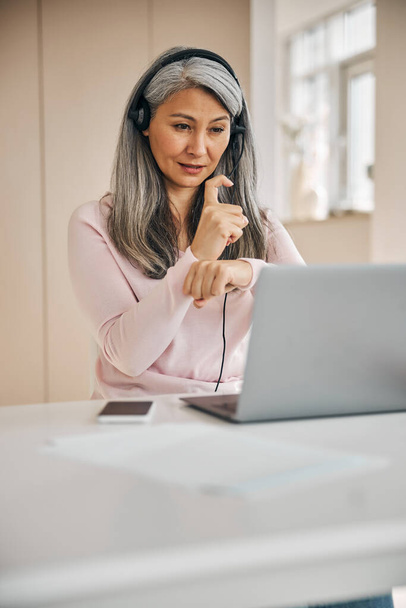 Mujer adulta concentrada en auriculares trabajando en línea con otras personas de la oficina en el hogar - Foto, Imagen