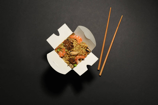 Box of wok noodles with seafood and chopsticks on black background, flat lay - Fotoğraf, Görsel