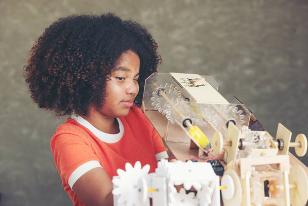 Poważny afro nastolatka dziewczyna kręcone włosy styl patrząc na części mechanizmu symulacyjnego robot model drewniany z podejrzeniem. - Zdjęcie, obraz