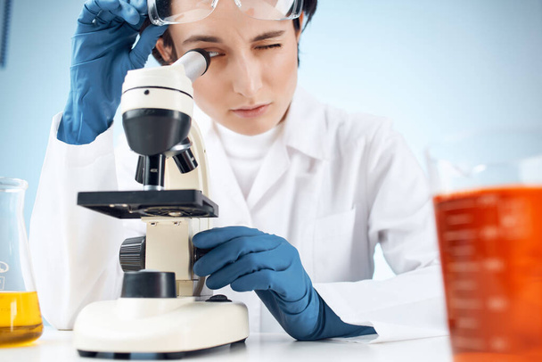 asistente de laboratorio en una máscara médica mirando a través de un microscopio  - Foto, Imagen