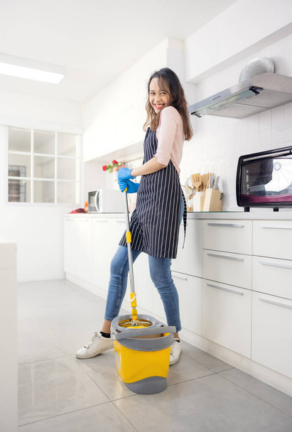 Портрет веселой молодой домохозяйки, держащей чистящие средства, улыбающейся и смотрящей в камеру на кухне. Профессиональный специалист по уборке, предлагающий помощь по хозяйству - Фото, изображение