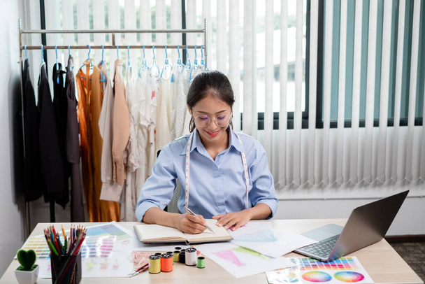 Hermosa joven sastre de moda asiática con accesorios de costura textil y bocetos de diseñador empresario están llenos de colores brillantes en el escritorio con un ordenador portátil para ideas creativas en el estudio. - Foto, Imagen