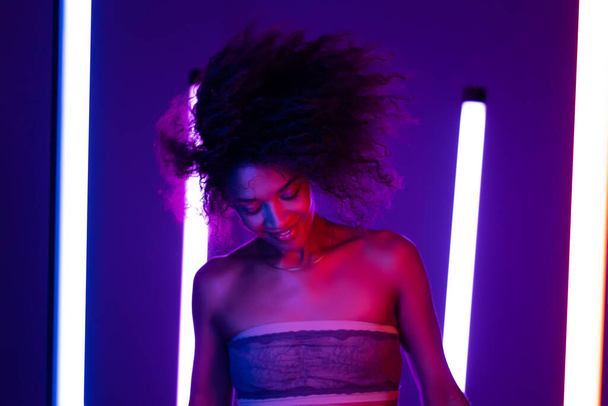 Multi ethnic woman with curly african hair dancing head on violet background in purple neon light. Sexy charming lady, night life concept. Modern pop outfit - bandeau - Foto, imagen