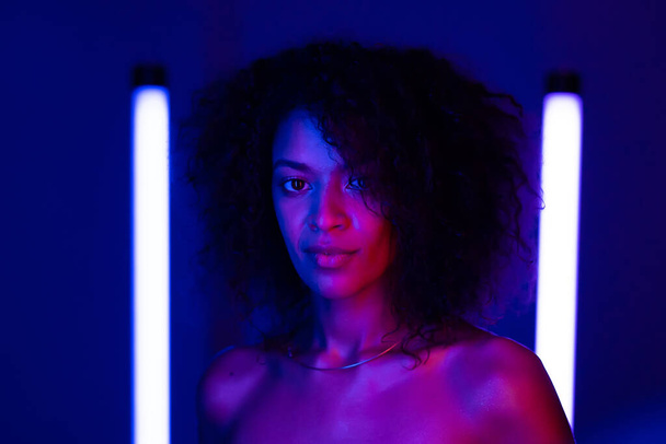Mixed race nude woman with curly african hair on blue background in pink neon light. Sexy lady without clothing looking at camera. - Foto, Bild