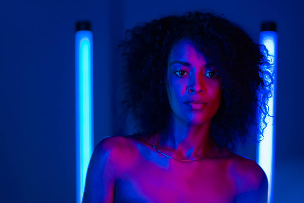 Mixed race nude woman with curly african hair on blue background in pink neon light. Sexy lady without clothing looking at camera. - Foto, afbeelding
