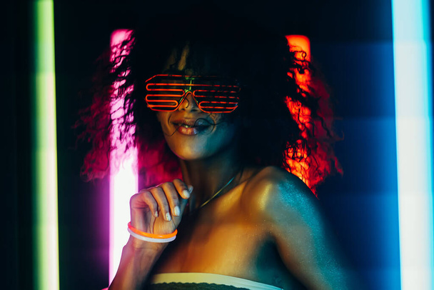 Cyberpunk style portrait of glamorous african woman on party under glowing colorful light. Nightclub, futuristic costume and neon glasses. Teenager, zoomer Z-generation. - Foto, Imagen