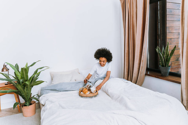 Afro-american girl resting in bed at home - Beautiful woman relaxing at home having breakfast in bed - Zdjęcie, obraz