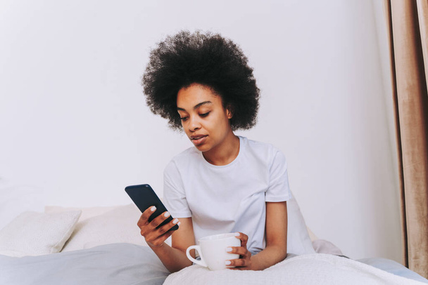 Afro-american girl resting in bed at home - Beautiful woman relaxing at home having breakfast in bed while checking her smartphone - Photo, Image