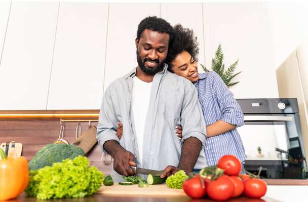 Beautiful afro american couple cooking at home - Beautiful and cheerful black couple preparing dinner together in the kitchen - Photo, Image