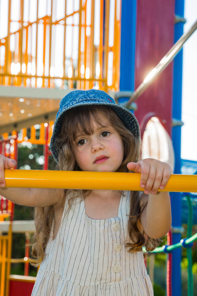  Attraction on the playground in green park. Warm summer day. Adorable little girl with long blonde hair in a white sundress and denim panama.  - Foto, immagini