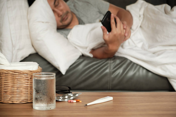 Asian man is absent from work and sick leave because of illness, takes temperature by thermometer, takes medicine, and sleeps in blankets on sofa at home, tissues, and capsule pills on the table. - Foto, immagini