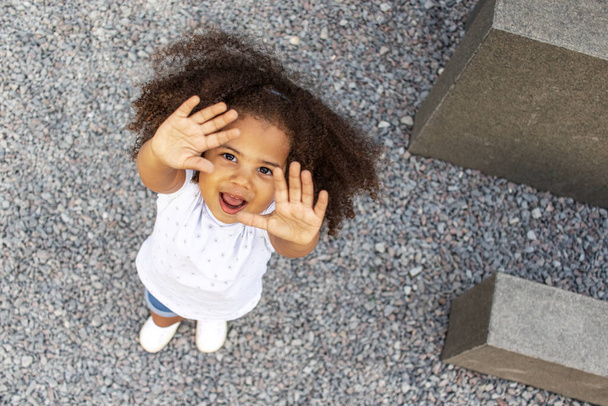 Outdoor portret van een krullend lachend Afrikaans Amerikaans meisje met opgeheven handen. Hoge hoekaanzicht. - Foto, afbeelding