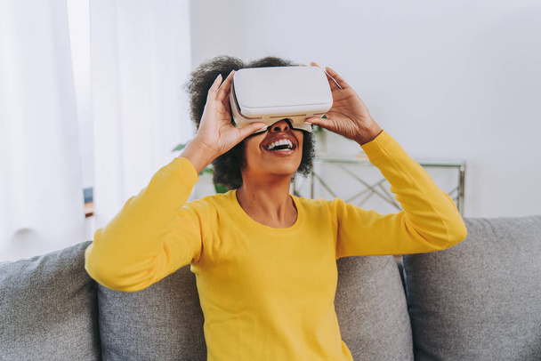 Beautiful woman playing at home with VR headset - Black woman lying down on sofa while interacting with augmented reality technology - Foto, Imagem