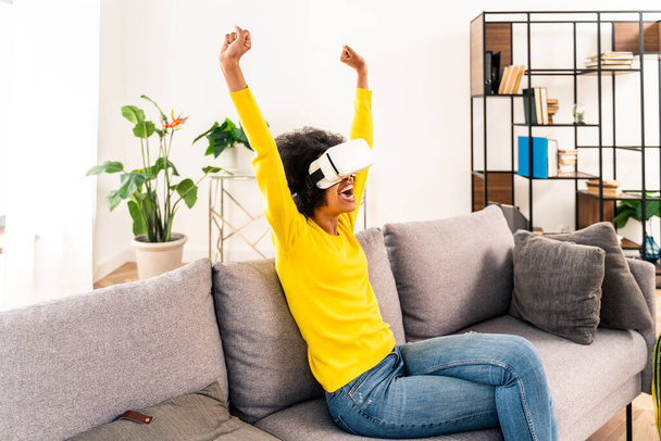 Beautiful woman playing at home with VR headset - Black woman lying down on sofa while interacting with augmented reality technology - Foto, imagen