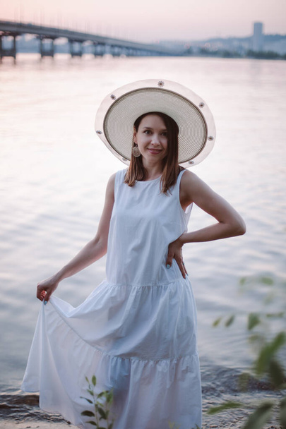 джентльменский портрет очаровательной кавказской женщины в белом летнем платье и соломенной шляпе, наслаждающейся нежными красками заката, молодая женщина отдыхает у реки на закате на свидании - Фото, изображение