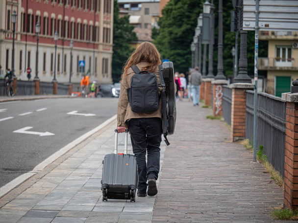 Menino de cabelos compridos visto por trás, andando em uma rua pública com uma mala de carrinho e uma guitarra nas costas. - Foto, Imagem