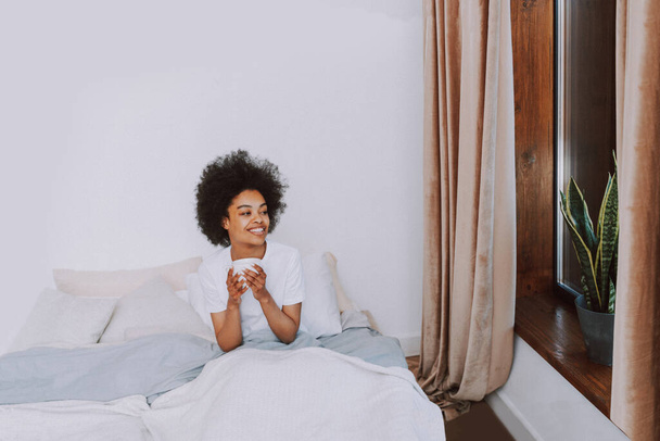 Afro-american girl resting in bed at home - Beautiful woman relaxing at home having breakfast in bed - Fotografie, Obrázek