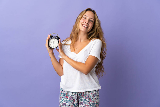 Mujer rubia joven aislada sobre fondo púrpura en pijama y celebración del reloj con expresión feliz - Foto, Imagen