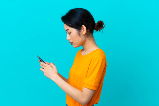 Nuori vietnamilainen nainen eristetty sinisellä taustalla lähettää viestin tai sähköpostia matkapuhelimella - Valokuva, kuva