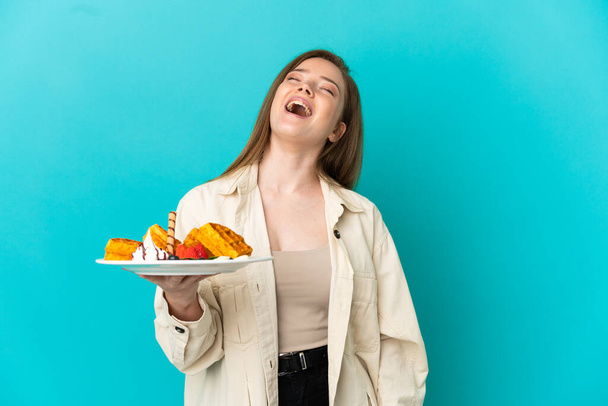 Девочка-подросток держит вафли на изолированном синем фоне, смеясь - Фото, изображение