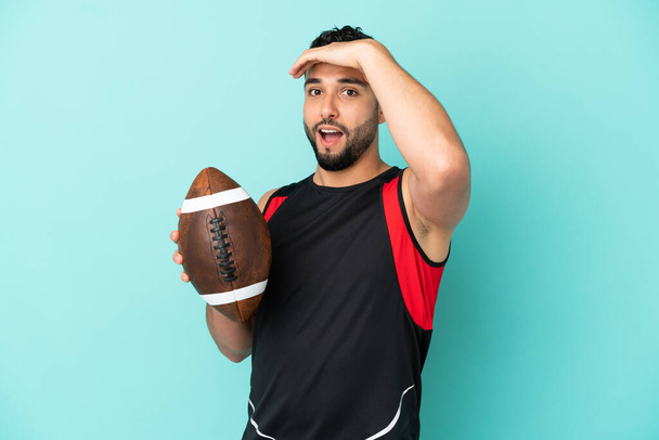 Young arab man playing rugby isolated on blue background doing surprise gesture while looking to the side - Photo, Image