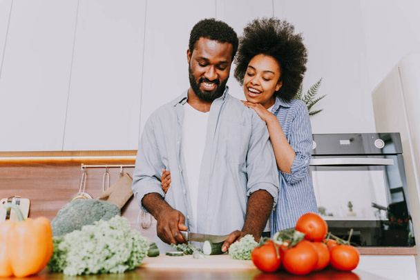 Beautiful afro american couple cooking at home - Beautiful and cheerful black couple preparing dinner together in the kitchen - Фото, изображение