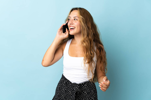 Young blonde woman isolated on blue background keeping a conversation with the mobile phone - Photo, image