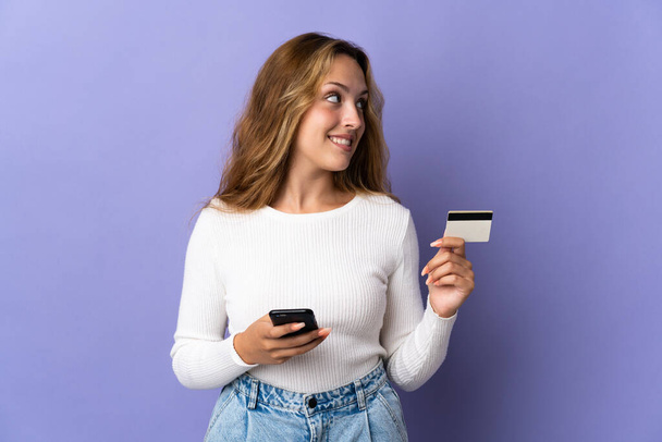 Молода блондинка ізольована на фіолетовому фоні, купуючи з мобільним телефоном з кредитною карткою, думаючи
 - Фото, зображення