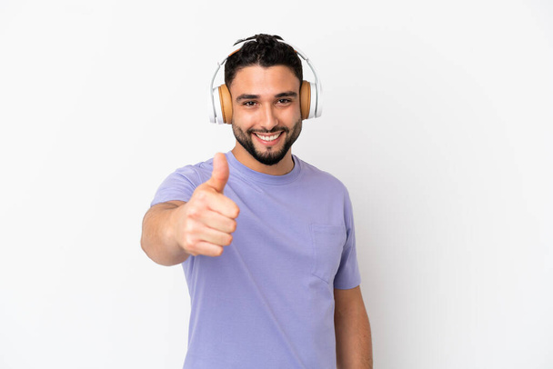 Young arab man isolated on white background listening music and with thumb up - Foto, Imagen