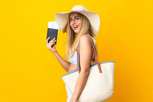 Young Uruguayan blonde woman in swimsuit holding a passport over isolated yellow background smiling a lot - Photo, Image