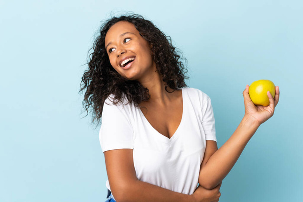 Teenager cuban girl isolated on blue background with an apple and happy - Φωτογραφία, εικόνα