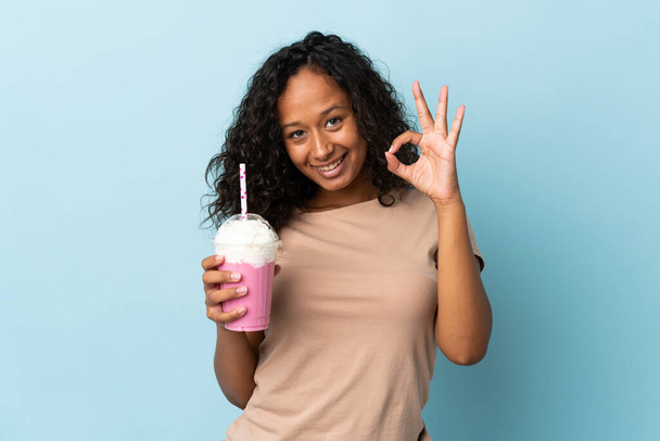 Chica adolescente con batido de fresa aislado sobre fondo azul mostrando signo de ok con los dedos - Foto, Imagen
