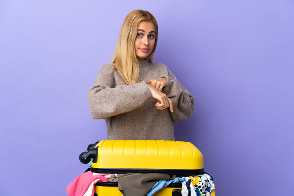 Молодая уругвайская блондинка с чемоданом одежды на изолированной фиолетовой стене делает жест опоздания - Фото, изображение