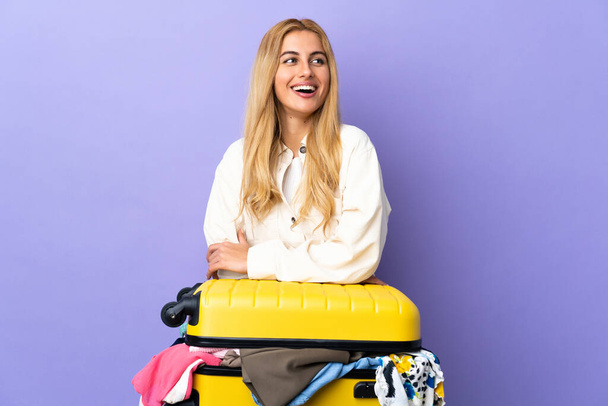 Mladá uruguayská blondýnka s kufrem plným šatů přes izolovanou fialovou stěnu šťastná a usmívající se - Fotografie, Obrázek