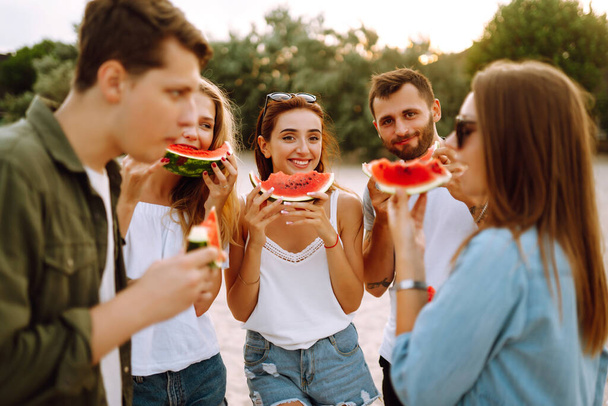 Young friends relaxing on the beach and eating watermelon. Group of young sitting together sitting near the sea and  enjoy summer party. People, lifestyle, travel, nature and vacations concept. - Φωτογραφία, εικόνα