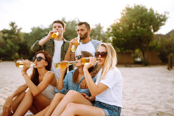 Group of friends cheers and drink beers on the beach. Young  friends sitting together sitting near the sea and enjoy summer party. Beach holiday and summer vacation concept. - Fotó, kép