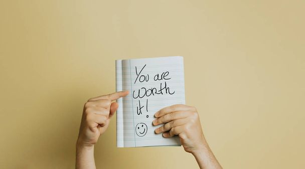 A person's hands holding an encouraging YOU ARE WORTH IT note written on paper against a yellow background - Foto, Imagen