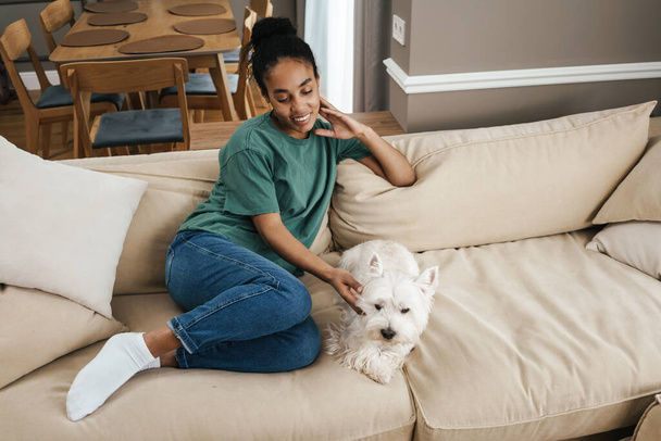 Smiling mid aged african woman sitting on a couch with her dog at home - Fotoğraf, Görsel
