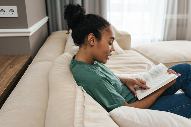 Young black woman reading book while sitting with her dog on sofa at home - Photo, Image