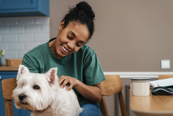 Smiling black woman stroking her dog while sitting at home kitchen - Foto, afbeelding