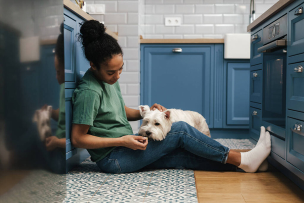 Smiling mid aged african woman playing with her dog in the kitchen, sitting on a floor - Foto, Bild