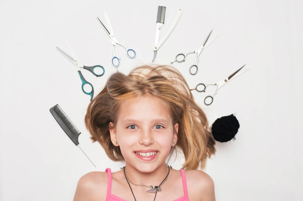 Das Mädchen liegt auf einem weißen Hintergrund zwischen professionellen Haarschneidewerkzeugen - Schere und Kamm liegen in der Nähe ihres Kopfes. Das Mädchen ist glücklich und lächelt, weil sie vor kurzem geschnitten und eine Frisur gemacht kare - Foto, Bild